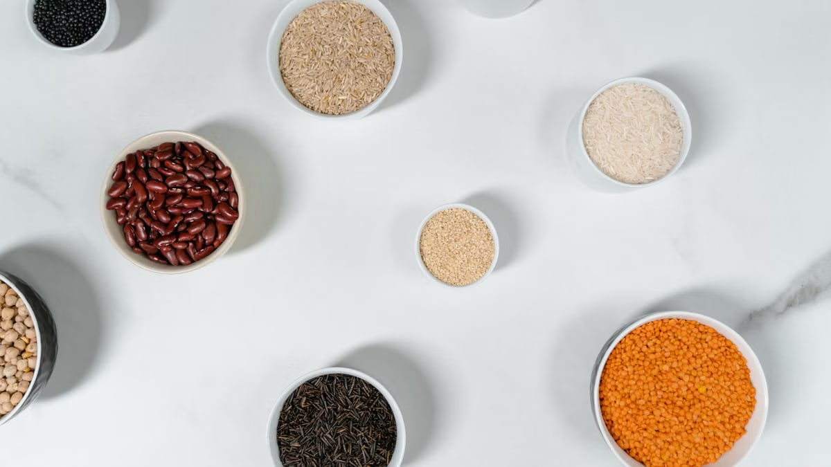 Top view of various grains and legumes in bowls on a white marble surface.